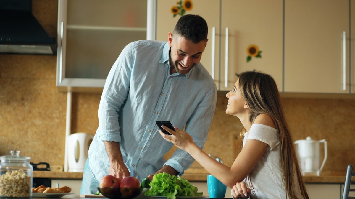 Attractive couple chatting in the kitchen early morning. Beautiful girl sharing social media on smartphone with her boyfriend cooking breakfast at home